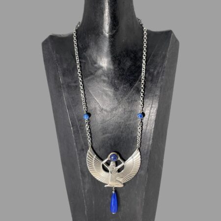 Main photo of Egyptian revival Isis necklace displayed on a black wooden stand with the necklace front forward facing. This stand is used for longer pieces and this necklace has a generous length with the goddess Isis taking centre stage of course! She is definitely a statement here She is kneeling with head and body facing right. Her arms are extended above her head where she holds a ball of lapis lazuli. The wings extend from behind the torso to the left and right and the tips of these are where the chain of the necklace is attached. 2 smaller lapis beads are added approximately 2 inches above the wing tips and then the chain continues all the way to the clasp. Below the goddess is a long water droplet shaped bead of lapis lazuli which hangs from the centre completing the look.