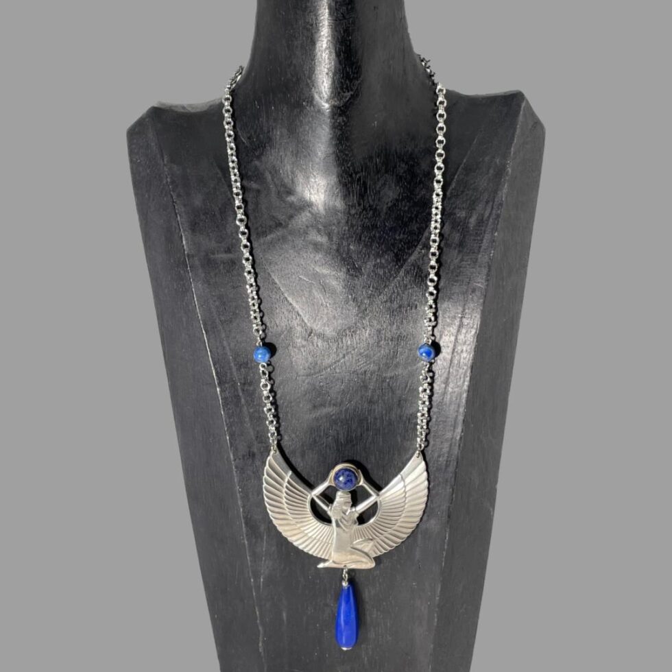 Main photo of Egyptian revival Isis necklace displayed on a black wooden stand with the necklace front forward facing. This stand is used for longer pieces and this necklace has a generous length with the goddess Isis taking centre stage of course! She is definitely a statement here She is kneeling with head and body facing right. Her arms are extended above her head where she holds a ball of lapis lazuli. The wings extend from behind the torso to the left and right and the tips of these are where the chain of the necklace is attached. 2 smaller lapis beads are added approximately 2 inches above the wing tips and then the chain continues all the way to the clasp. Below the goddess is a long water droplet shaped bead of lapis lazuli which hangs from the centre completing the look.