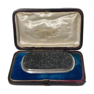 Main photo of antique tourmaline specimen brooch displayed inside its fitted case. The brooch is a wide oblong shape bordering on rectangular. The tourmaline is a grey/black colour and in a natural crystalline form and not polished smooth. The silver frame around the tourmaline is relatively thin in comparison but nicely executed with a plain flat silver frame around the edge of the tourmaline followed by a twisted silver band and finally set on a smooth round edged silver main frame. This brooch is all about the tourmaline! Black tourmaline is renowned for grounding the mind, relieving stress and shielding from negativity. Owing to its size and weight, this brooch would ideally suit a heavy overcoat or jacket.