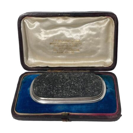 Main photo of antique tourmaline specimen brooch displayed inside its fitted case. The brooch is a wide oblong shape bordering on rectangular. The tourmaline is a grey/black colour and in a natural crystalline form and not polished smooth. The silver frame around the tourmaline is relatively thin in comparison but nicely executed with a plain flat silver frame around the edge of the tourmaline followed by a twisted silver band and finally set on a smooth round edged silver main frame. This brooch is all about the tourmaline! Black tourmaline is renowned for grounding the mind, relieving stress and shielding from negativity. Owing to its size and weight, this brooch would ideally suit a heavy overcoat or jacket.