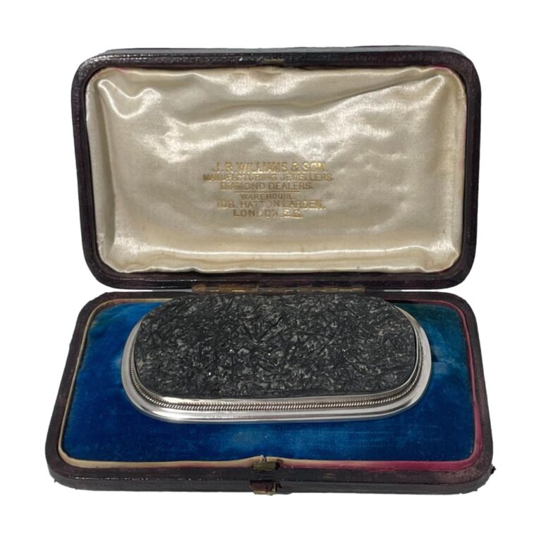 Main photo of antique tourmaline specimen brooch displayed inside its fitted case. The brooch is a wide oblong shape bordering on rectangular. The tourmaline is a grey/black colour and in a natural crystalline form and not polished smooth. The silver frame around the tourmaline is relatively thin in comparison but nicely executed with a plain flat silver frame around the edge of the tourmaline followed by a twisted silver band and finally set on a smooth round edged silver main frame. This brooch is all about the tourmaline! Black tourmaline is renowned for grounding the mind, relieving stress and shielding from negativity. Owing to its size and weight, this brooch would ideally suit a heavy overcoat or jacket.