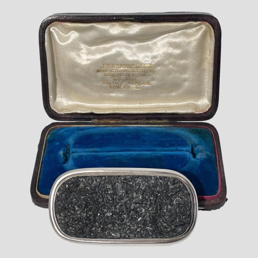 Photo of the specimen tourmaline brooch displayed outside of its open case and propped up in front forward facing. This is to show the fitted section for the brooch in the vibrant blue velvet. The logo in gold for jewellers J. R. Williams can be seen on the pearly coloured silk inside the lid. The interior of the case is in fairly good condition with minor wear.