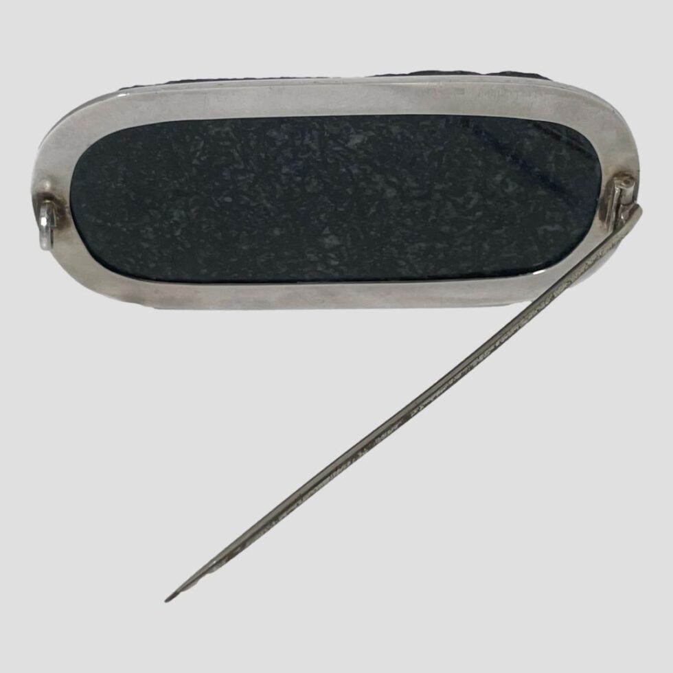 Photo of the back of the tourmaline brooch showing the silver setting. The brooch is propped up by its own pin which is shown open. The silver frame lies flat against the stone which has been smoothed to fit securely to the silver.