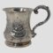 Main photo of Victorian sterling silver cup seen from an eye level with the handle to the right. On the side, there is a boat like shaped window with foliage below and a few flowers with a single leaf above. To the right just below the rim you can see the hallmark. The handle is shaped like half an outline of a heart-curving upwards from the tip of the rim, out in a curve then tapering and attaching to the lower part of the cup.