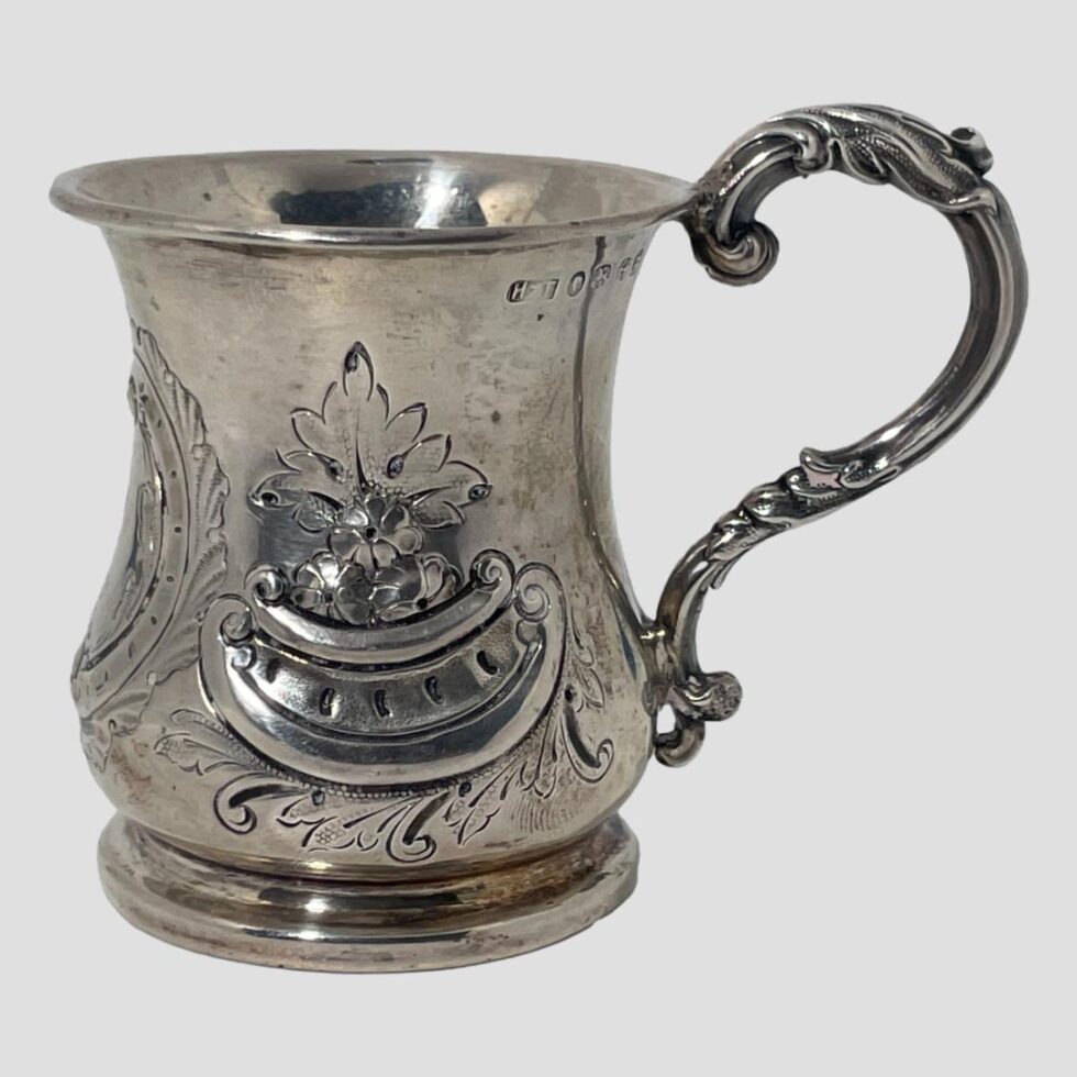 Main photo of Victorian sterling silver cup seen from an eye level with the handle to the right. On the side, there is a boat like shaped window with foliage below and a few flowers with a single leaf above. To the right just below the rim you can see the hallmark. The handle is shaped like half an outline of a heart-curving upwards from the tip of the rim, out in a curve then tapering and attaching to the lower part of the cup.