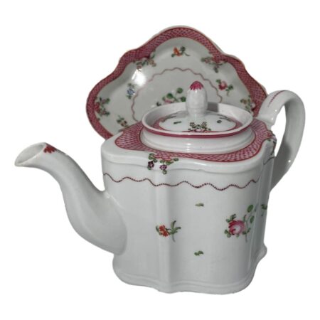 Main photo of New Hall teapot &stand shown with the stand displayed upright behind the teapot. The teapot spout is facing a little towards the bottom left corner. New Hall were pioneers in hard paste porcelain clearly a design quality that withstood the test of time. The base colour is white with minimal floral design mostly in pink to the main body. Around the outer rim at the top, there is a band of pink netting like pattern which continues around the outside of the teapot lid. The stand, which would be mostly obscured by the teapot, almost mirrors the teapot design pattern.