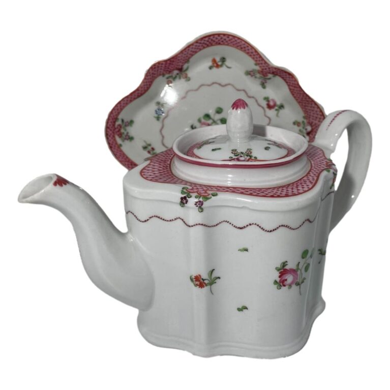 Main photo of New Hall teapot &stand shown with the stand displayed upright behind the teapot. The teapot spout is facing a little towards the bottom left corner. New Hall were pioneers in hard paste porcelain clearly a design quality that withstood the test of time. The base colour is white with minimal floral design mostly in pink to the main body. Around the outer rim at the top, there is a band of pink netting like pattern which continues around the outside of the teapot lid. The stand, which would be mostly obscured by the teapot, almost mirrors the teapot design pattern.