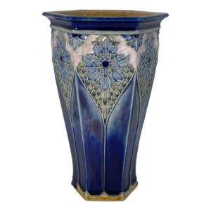Main photo of Royal Doulton hexagonal vase see with one flat edge in the centre foreground with half of others on left and right visible. The base colour of the vase is a rich deep blue, not dark but with depth. The base rim and inside rim of mouth is a mustard yellow glaze. The hexagonal shape tapers at the base with cathedral window like forms rising up from the base rim pointed at the top. Here, we have a stunning floral pattern almost like stained glass mostly in blues and green with a pale pink section just above the large 'stained glass' flower. The outer rim of the vase is the rich deep blue again with small half flowers evenly spaced out and each hexagon edge. Really a very stunning and unusual design piece.