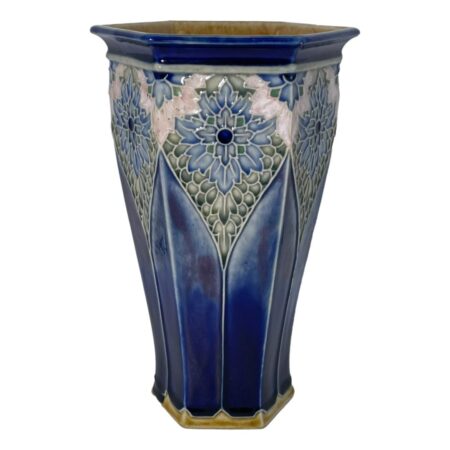 Main photo of Royal Doulton hexagonal vase see with one flat edge in the centre foreground with half of others on left and right visible. The base colour of the vase is a rich deep blue, not dark but with depth. The base rim and inside rim of mouth is a mustard yellow glaze. The hexagonal shape tapers at the base with cathedral window like forms rising up from the base rim pointed at the top. Here, we have a stunning floral pattern almost like stained glass mostly in blues and green with a pale pink section just above the large 'stained glass' flower. The outer rim of the vase is the rich deep blue again with small half flowers evenly spaced out and each hexagon edge. Really a very stunning and unusual design piece.