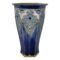 Main photo of Royal Doulton hexagonal vase see with one flat edge in the centre foreground with half of others on left and right visible. The base colour of the vase is a rich deep blue, not dark but with depth. The base rim and inside rim of mouth is a mustard yellow glaze. The hexagonal shape tapers at the base with cathedral window like forms rising up from the base rim pointed at the top. Here, we have a stunning floral pattern almost like stained glass mostly in blues and green with a pale pink section just above the large 'stained glass' flower. The outer rim of the vase is the rich deep blue again with small half flowers evenly spaced out and each hexagon edge. Really a very stunning and unusual design piece.
