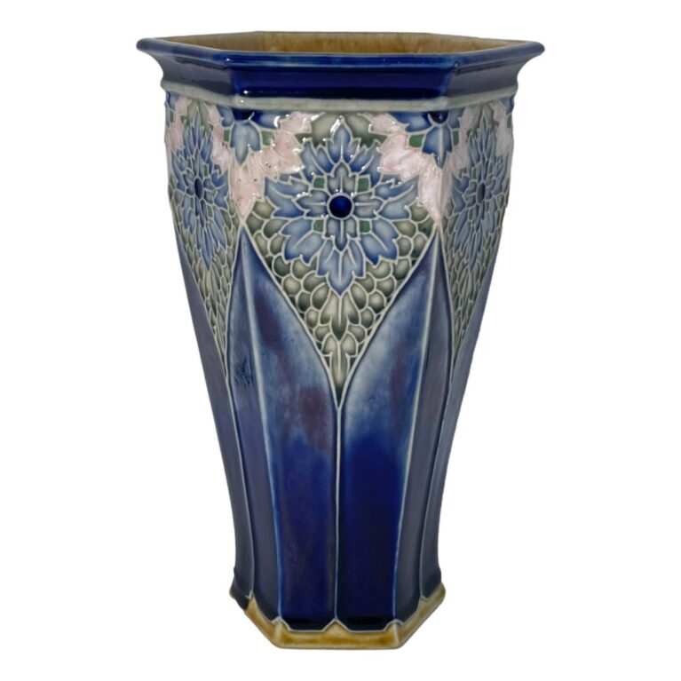 Main photo of Royal Doulton hexagonal vase see with one flat edge in the centre foreground with half of others on left and right visible. The base colour of the vase is a rich deep blue, not dark but with depth. The base rim and inside rim of mouth is a mustard yellow glaze. The hexagonal shape tapers at the base with cathedral window like forms rising up from the base rim pointed at the top. Here, we have a stunning floral pattern almost like stained glass mostly in blues and green with a pale pink section just above the large 'stained glass' flower. The outer rim of the vase is the rich deep blue again with small half flowers evenly spaced out and each hexagon edge. Really a very stunning and unusual design piece.