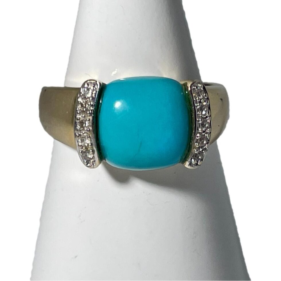 Main photo of 14kt turquoise diamond ring displayed on a white cone shaped stand and forward facing. The turquoise is almost flawless with just a small dark line inclusion. On either side are thin columns that run alongside the curve of the cushion cut turquoise. Each column has 5 round cut diamonds. The 14 karat gold band is solid and the shoulders are almost the width of the turquoise, tapering to a slightly thinner band at the back.