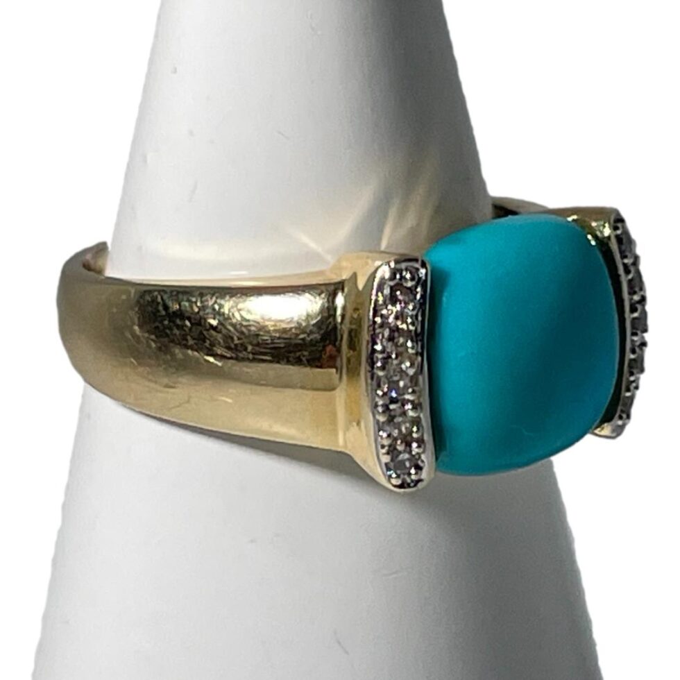 Photo of gold turquoise ring still displayed on a white cone shaped stand this time with the ring front facing towards the bottom right corner showing the solid and thick shoulder of the band.