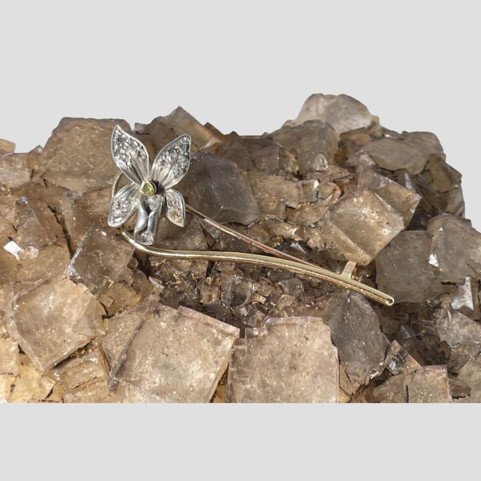 Main photo of Edwardian diamond orchid brooch displayed on top of a rock of natural river quartz. The orchid flower is made of silver with diamonds in its 2 petals, 2 sepals and its lip or labellum. The column is accentuated with a peridot stone which has its own gold casing. The stem of the flower curves out and round from the side of the flower head and passes below it ending at the extreme right. The pin follows the route of the stem.