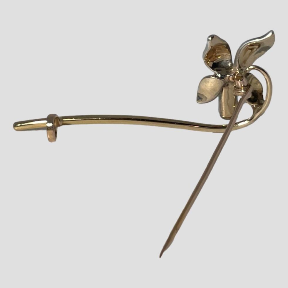 Photo of the back of the orchid flower brooch showing the pin open and holding the brooch upright. The back of the orchid flower is in gold and the curved design of the stem is visible here.