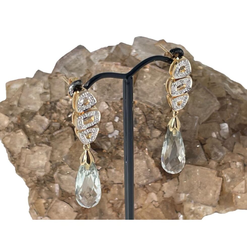 Photo of aquamarine drop and diamond earrings displayed on a black display stand with a rock of natural river quartz behind. The quartz is a pink sand colour which brings out the subtle blue hue of the aquamarine stones. The earring fronts are facing to the bottom right and we are looking from a slight height down at the earrings.