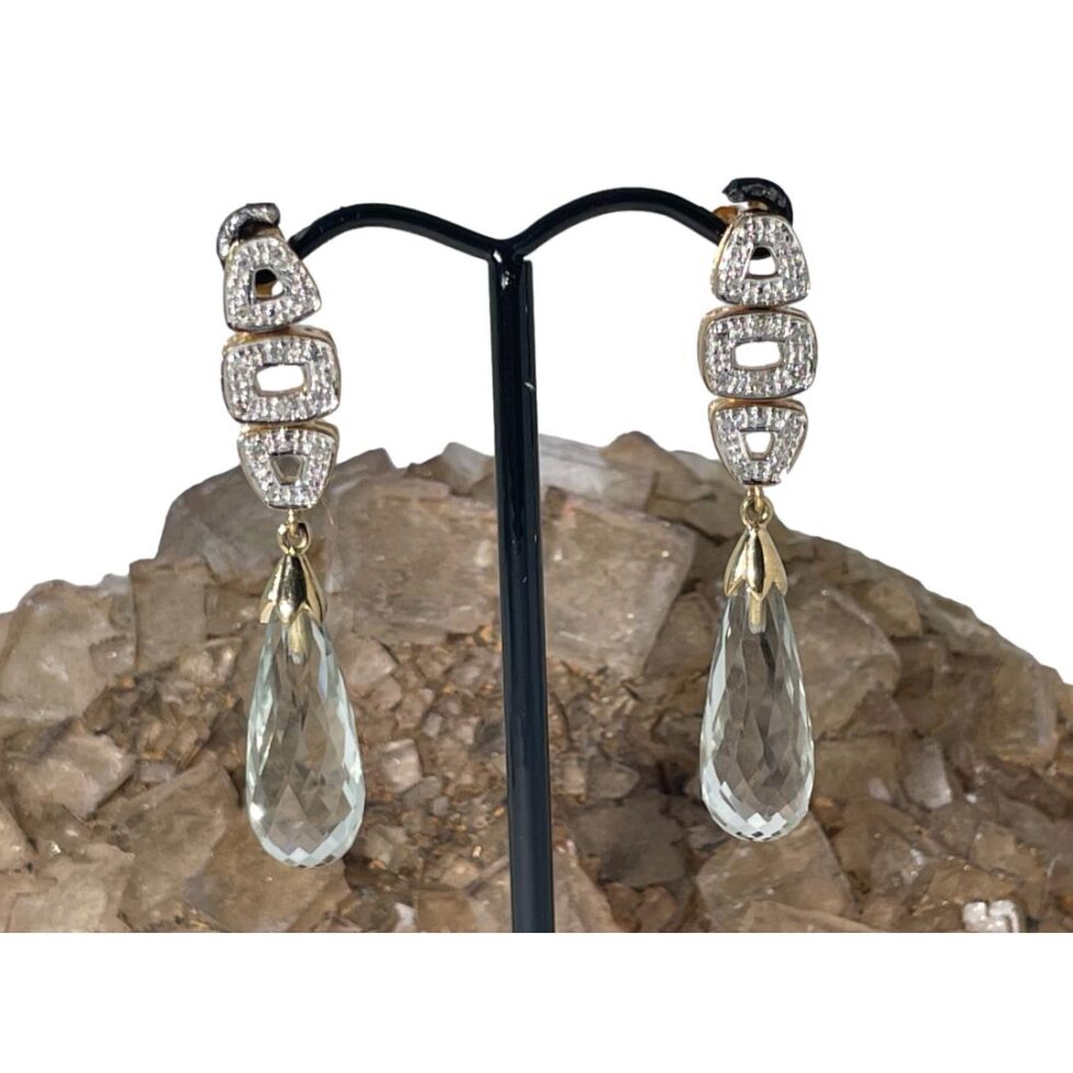 Main photo of gold aquamarine diamond earrings displayed hanging from a black stand and forward facing. The top part of the earrings housing the triple sectioned diamond studded column is against a white background whilst the aquamarine droplets have the river quartz as a backdrop. The stones are a subtle pale blue colour which becomes clearer against the quartz.