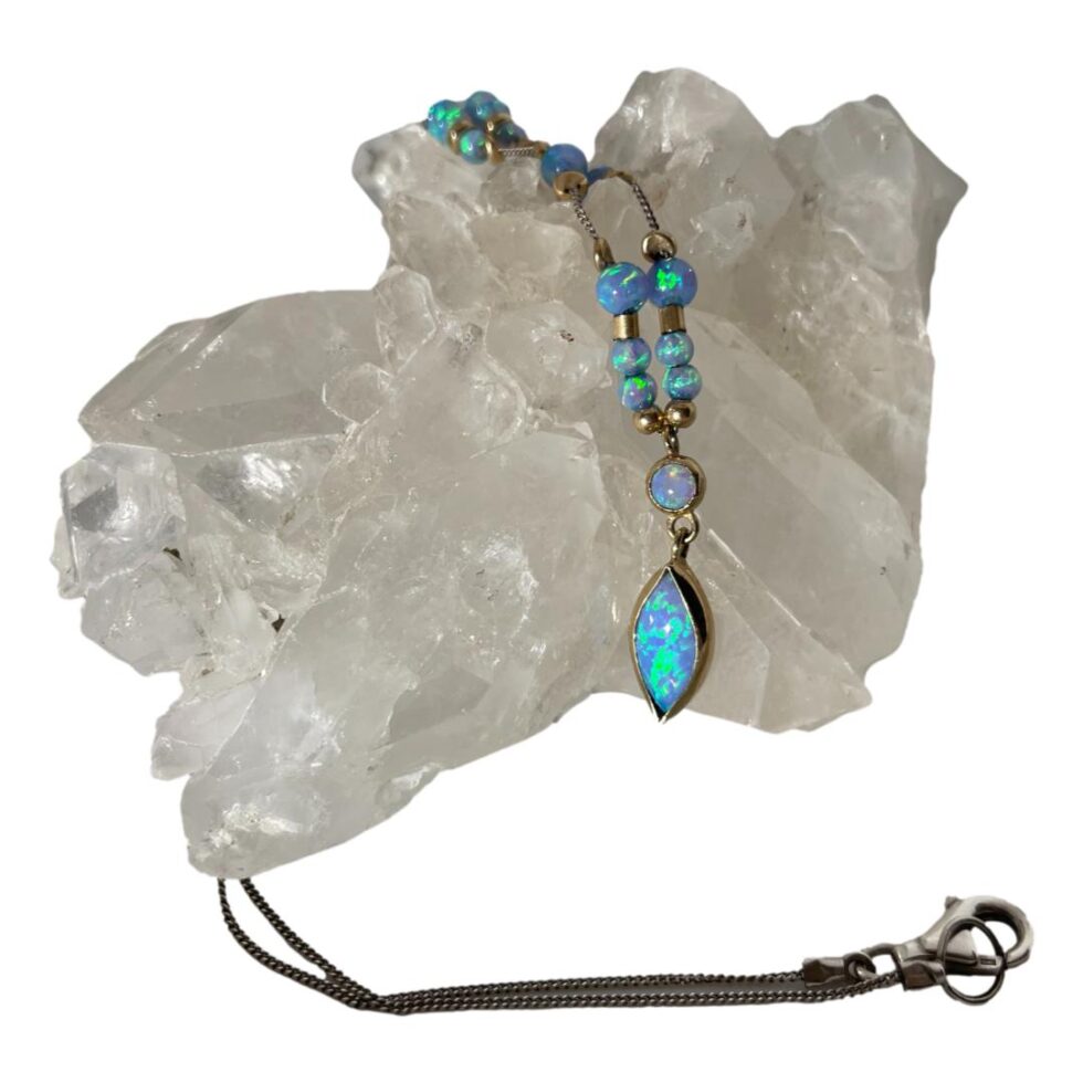 Main photo of opal gold silver necklace displayed draped across a rock of natural quartz crystal with the opal side hanging from the rock and the silver chain coming around the back and across the front. The small attached pendant at the very front has a vertical almond shaped stone encased in a 9 karat gold frame and a smaller round opal cabochon also encased in its own gold frame above. This short pendant is then attached to the silver chain which is barely visible here as there are gold divider beads separating the black opal beads. From the pendant area, there are 2 gold ball beads followed by 2 small black opal beads. Next is a short gold tube divider bead then a larger round opal bead. Next divider bead is an upright flat round and finally we find a bit of silver chain appearing. There are another 3 sections with gold divider beads and opals in different sequence then silver chain and clasp to finish. Nice thing about this necklace is that the main parts with the black opal and gold beads are at the front. The black opal has a beautiful lagoon blue and turquoise base colour with flecks of green and yellow and hints of pink and orange.