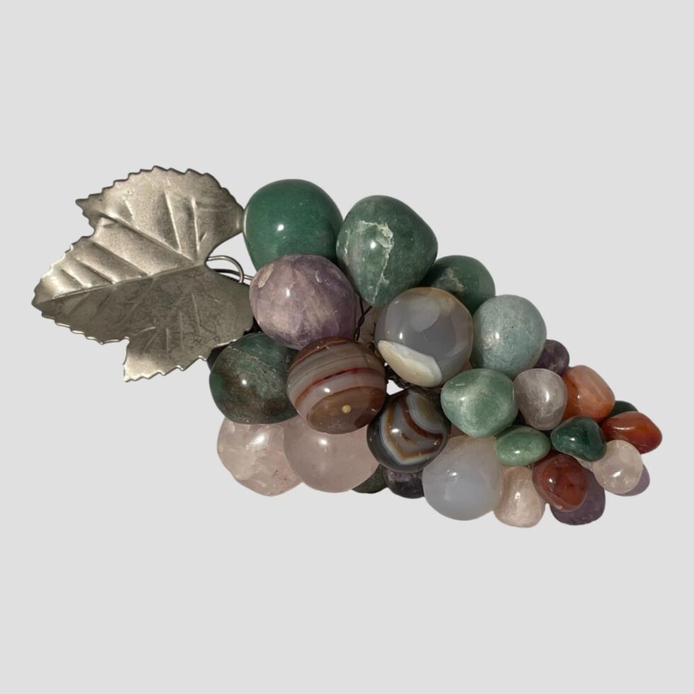 Main photo of vintage semi-precious stone grapes shown laid horizontally across the page with the top of the bunch on the left. The grapes are made of different semi-precious stones including amethyst, rose quartz, aventurine, agate and more. Colours range from greens to purples, pinks and reds. There is a silver tone metal leaf and tendrils. Lovely decorative item making a great addition to faux fruit bowl.