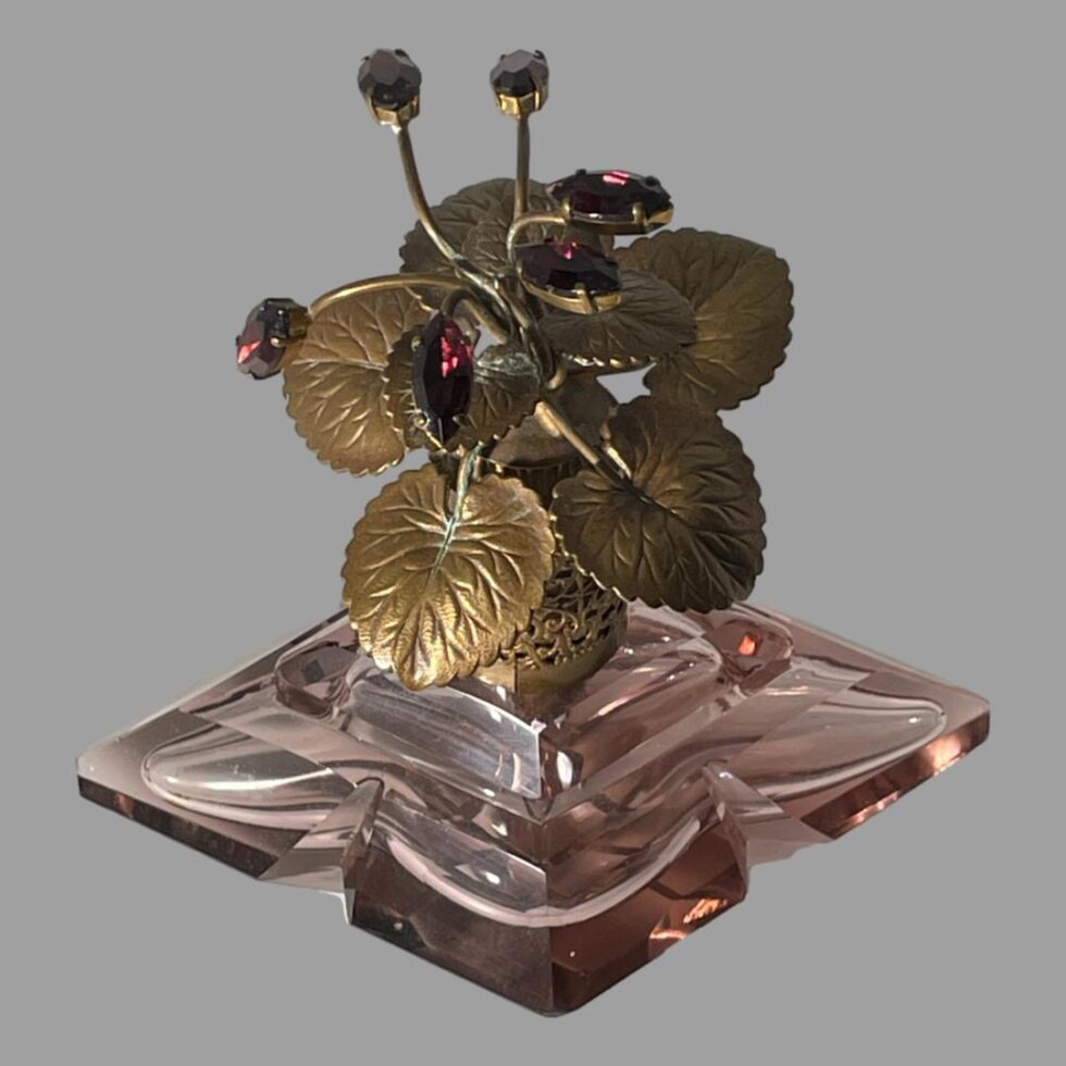 Photo of vintage scent bottle looking diagonally down from above. The wide side of the smoky lilac glass bottle is in the foreground and some of the purple rhinestone flower buds are visible giving a nice contrasting outlook of the 2 purple hues of this piece.