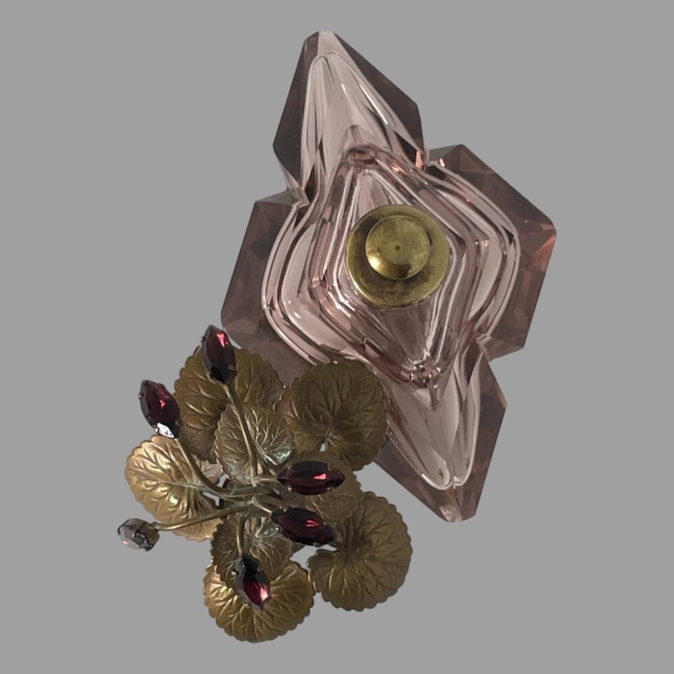 Photo looking straight down at the scent bottle from above. It has had its gilt metal leaf top removed and place to the bottom left. The brass pump nozzle top is visible in the centre of the diamond shaped cut glass bottle.