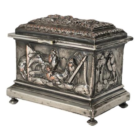 Main photo of French repousse jewellery casket displayed from a near eye level with the front opening side facing towards the bottom left and one side visible facing the bottom right corner. The casket sits on 4 feet which appear quite dainty for the solid and heavily embossed (repousse) casket. The base and top of lid jut out a little over the main body of the casket. The scenes on the sides show a lone bird, possible a peacock upright and looking towards the other side which shows a cockerel with 2 hens in a yard with a prowling fox on the very left. Some of the silver plate has worn off and the copper hue is coming through on the hens breasts and a small patch on the head of the cockerel and fox. This wear and copper exposure adds extra depth & charm to the casket.