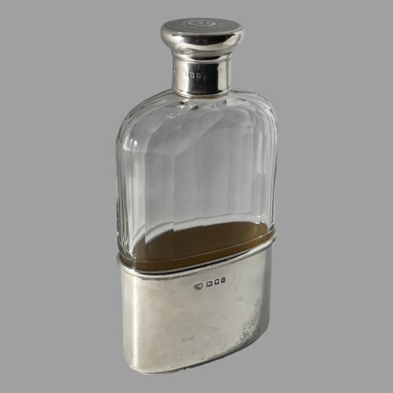 Main photo of c1918 silver hip flask displayed from an almost eye level just slightly raised. The flask is displayed at a slight angle with the hallmark visible to the silver cup at base and the neck of the screw top. The shoulders of the flask are turned to just off bottom left and top right corners.