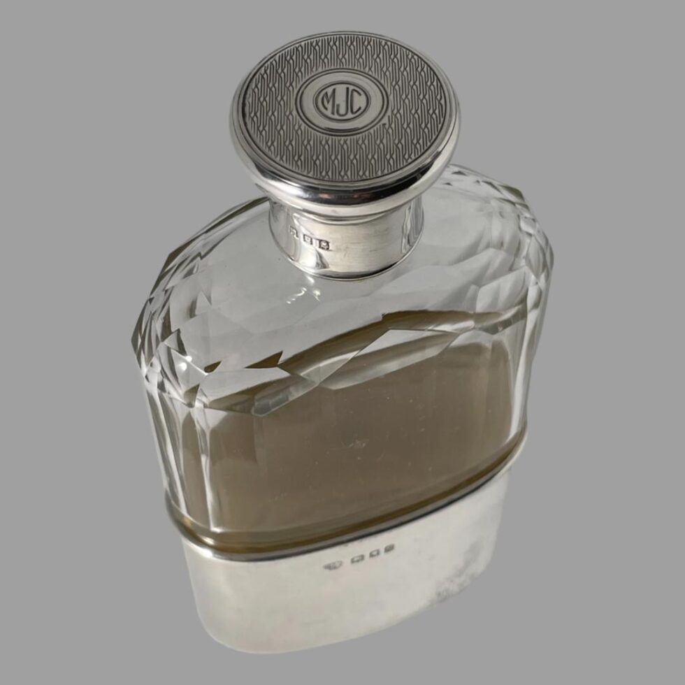 Photo looking diagonally down at the top of the hip flask which is displayed diagonally across the page with one flat side facing to the bottom right corner. The top of the engine turned lid with initials MJC at centre is clearly visible as is the lovely faceted detail of the glass flask.