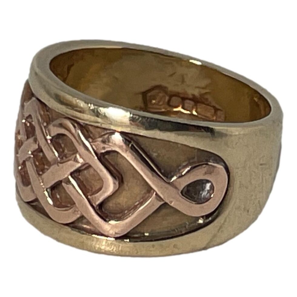 Photo of Clogau ring on a plain white surface with the ring front faced towards the bottom left. The hallmark on inside band is just visible.