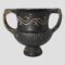 Main photo of small Romano-Greek urn seen from an eye level with the handles to the left and right. This lovely little urn is matt black in colour with a little decorative detail at the top of urn body. It stands on a single small round foot with a vertically ridged lower section. The handles extend from the top of this ridged area. The top section of urn is smooth with the decorative design which look like stylised waves. There is a slightly fluted lip at the mouth which the top of the handles attach to. These handles have the knotted or snake head ends attached.