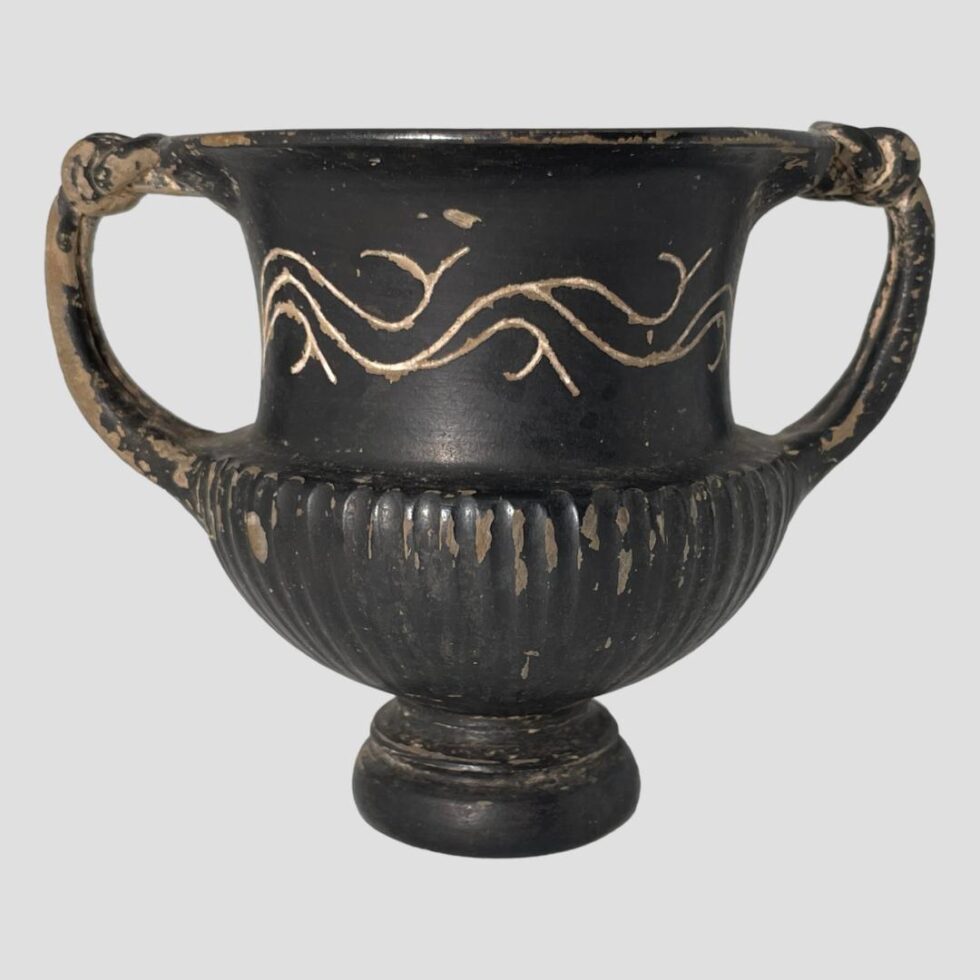 Main photo of small Romano-Greek urn seen from an eye level with the handles to the left and right. This lovely little urn is matt black in colour with a little decorative detail at the top of urn body. It stands on a single small round foot with a vertically ridged lower section. The handles extend from the top of this ridged area. The top section of urn is smooth with the decorative design which look like stylised waves. There is a slightly fluted lip at the mouth which the top of the handles attach to. These handles have the knotted or snake head ends attached.
