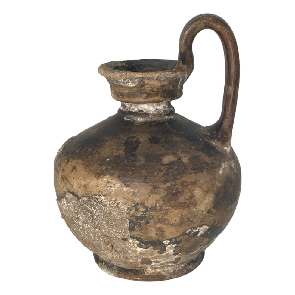 Main photo of Roman shipwreck wine jug displayed on a plain white background and seen from an eye level. The jug has a bulbous lower body for the wine, a short neck and a rounded mouth. The single handle is attached to one end of the mouth, rises upward then curves straight down to sit above the upper bulbous area of body. The jug has a lovely patina with some fossilisation along the bottom.