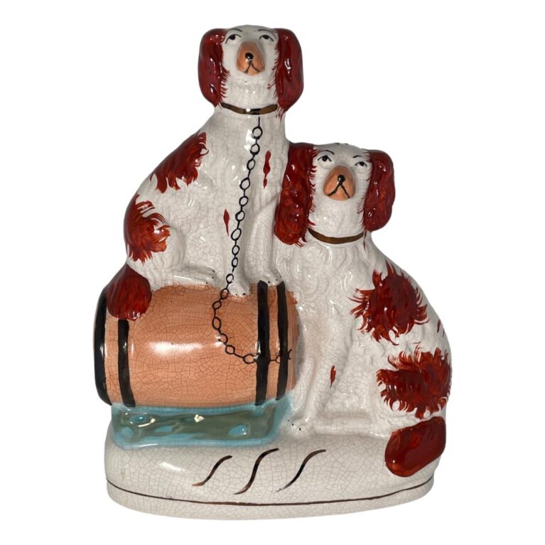 Main photo of Victorian Staffordshire Russet Spaniels displayed against a white background and shown forward facing. On the left, the spaniel is sat on top of the barrel and a chain is attached from its collar to the barrel lid (on the right) The barrel is itself sat on top of a blue cushion (?) On the immediate right of the barrel is the larger of the russet spaniels who is sat with body facing left towards the barrel. Both dogs are sat facing each other with heads only turned to camera. This is an eye level shot and at this angle the dogs appear to have a 'Not taking any of your nonsense' expression. They are serious at their guarding duties! Both dogs are white with russet patches.