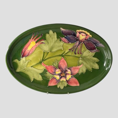 Main photo of Moorcroft Columbine dish displayed upright on a plate stand and forward facing. The base colour for the dish is the iconic Moorcroft green with light green leaves of the columbine across the central length of the dish. To the top left is a columbine flower in bud waiting to burst forth in pale red with yellow centre and on the top right is one in full bloom and seen from a side angle in deep mauve and mustard hues. At the centre bottom is another flower in full bloom and we are looking straight at it from the top in pale red with yellow centre and a touch of deep mauve within.