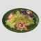 Main photo of Moorcroft Columbine dish displayed upright on a plate stand and forward facing. The base colour for the dish is the iconic Moorcroft green with light green leaves of the columbine across the central length of the dish. To the top left is a columbine flower in bud waiting to burst forth in pale red with yellow centre and on the top right is one in full bloom and seen from a side angle in deep mauve and mustard hues. At the centre bottom is another flower in full bloom and we are looking straight at it from the top in pale red with yellow centre and a touch of deep mauve within.