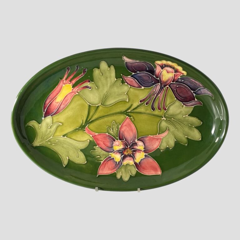 Main photo of Moorcroft Columbine dish displayed upright on a plate stand and forward facing. The base colour for the dish is the iconic Moorcroft green with light green leaves of the columbine across the central length of the dish. To the top left is a columbine flower in bud waiting to burst forth in pale red with yellow centre and on the top right is one in full bloom and seen from a side angle in deep mauve and mustard hues. At the centre bottom is another flower in full bloom and we are looking straight at it from the top in pale red with yellow centre and a touch of deep mauve within.