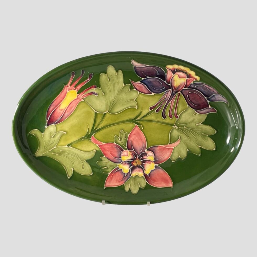 Main photo of Moorcroft Columbine dish displayed upright on a plate stand and forward facing. The base colour for the dish is the iconic Moorcroft green with light green leaves of the columbine across the central length of the dish. To the top left is a columbine flower in bud waiting to burst forth in pale red with yellow centre and on the top right is one in full bloom and seen from a side angle in deep mauve and mustard hues. At the centre bottom is another flower in full bloom and we are looking straight at it from the top in pale red with yellow centre and a touch of deep mauve within.