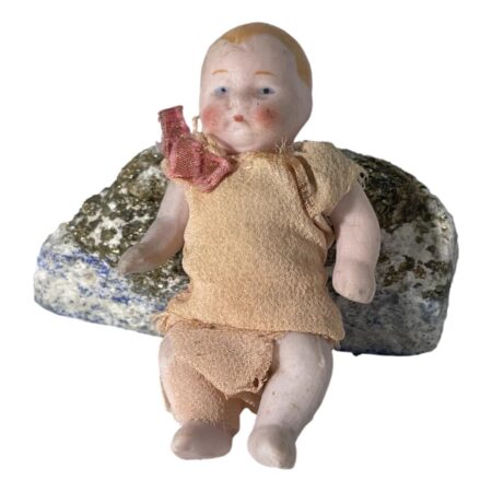 Main photo of miniature German bisque doll displayed leaning against a small rock and forward facing. The doll has painted ginger hair and eyebrows and blue eyes, rosy cheeks and a small red mouth. A fair amount of grubbiness is dotted around its face like its had a really good time playing in the dirt (we've all been there!) The clothes are in quite a poor state. In pale orange, it is like a fine muslin cloth which has clearly not stood the test of time. The pants are literally just hanging on while the top is in slighter better condition. There is a red bow visible to the right shoulder.