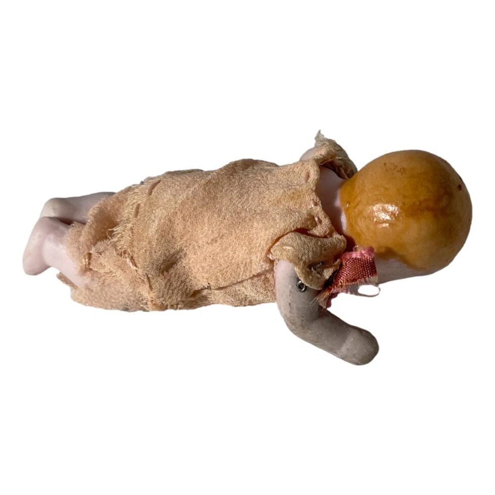 Photo of the bisque miniature doll now face down as if crawling. The painted ginger (orange) hair on its head is visible. The doll is facing to the right with feet on the left.