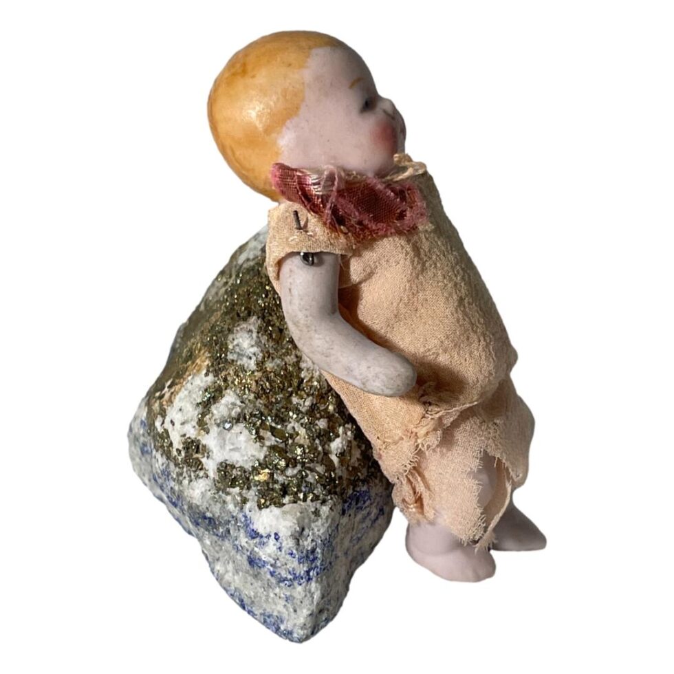Photo of the German miniature bisque doll still displayed leaning against a rock but this time from a side angle with its right arm in the foreground. The arm is quite grubby but also there is a visible sticking out through the shoulder of its muslin cloth top.