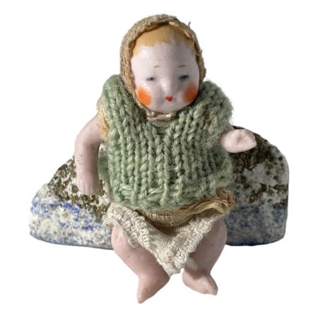 Main photo of German miniature bisque doll displayed leaning against a small rock and forward facing. This little doll is wearing a lacy cap and has pale orange hair painted underneath. It has more of a glossy face with round orange cheeks and small dark orange mouth. The woolly vest appears to be a later addition. Underneath the vest, it is wearing a top similar in design and material to the cap and it wears a pair of knitted short pants. Its left arm is made bent at the elbow.