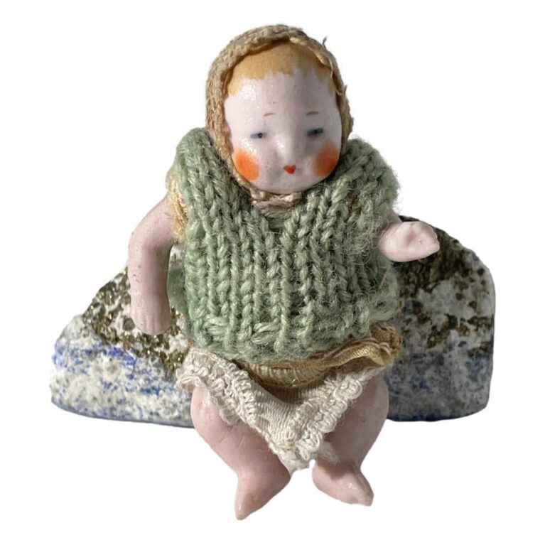 Main photo of German miniature bisque doll displayed leaning against a small rock and forward facing. This little doll is wearing a lacy cap and has pale orange hair painted underneath. It has more of a glossy face with round orange cheeks and small dark orange mouth. The woolly vest appears to be a later addition. Underneath the vest, it is wearing a top similar in design and material to the cap and it wears a pair of knitted short pants. Its left arm is made bent at the elbow.