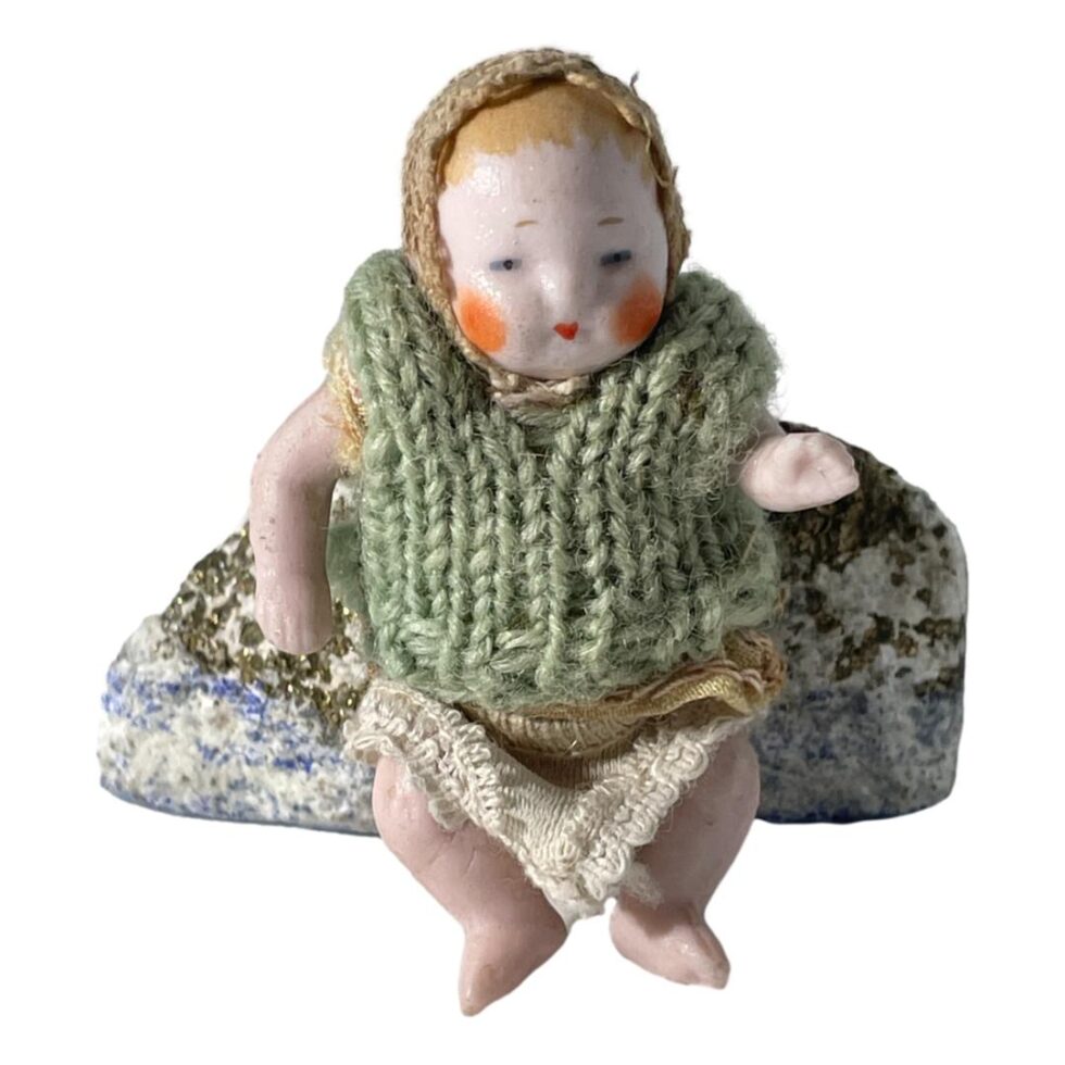 Main photo of German miniature bisque doll displayed leaning against a small rock and forward facing. This little doll is wearing a lacy cap and has pale orange hair painted underneath. It has more of a glossy face with round orange cheeks and small dark orange mouth. The woolly vest appears to be a later addition. Underneath the vest, it is wearing a top similar in design and material to the cap and it wears a pair of knitted short pants. Its left arm is made bent at the elbow.
