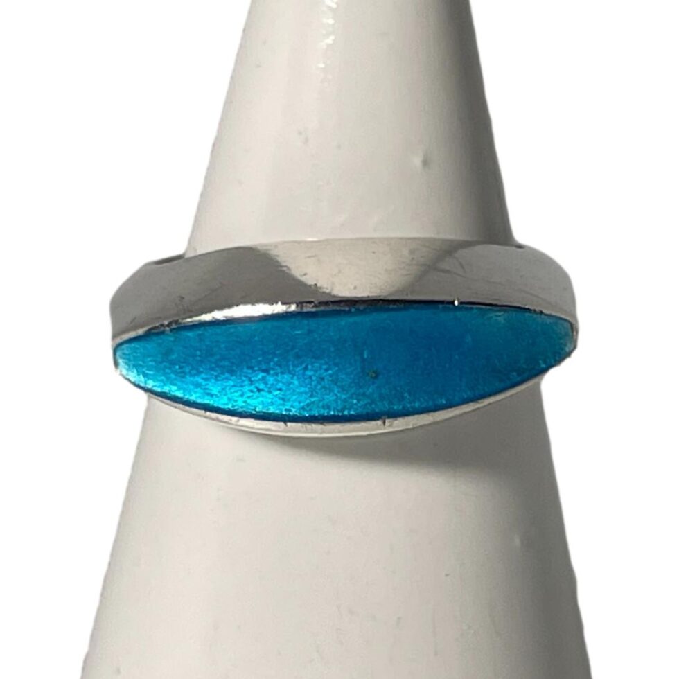 Main photo of Just Andersen enamel ring displayed on a white cone shaped stand with the ring front forward facing. The navette shaped turquoise blue enamel is very eye catching with the flaw in one end barely visible. The ring has quite a wide gallery in solid silver with the enamel sitting just at the front. A vert sleek, stylish and stunning ring!