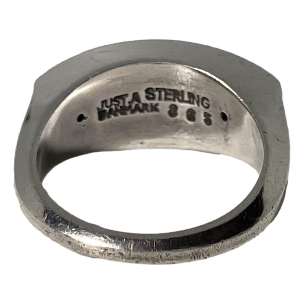 Photo of ring on a flat surface and from the back band. The hallmark is debossed to the back of the ring front and reads Just A Danmark to the left, Sterling to the right and number 363 below sterling.