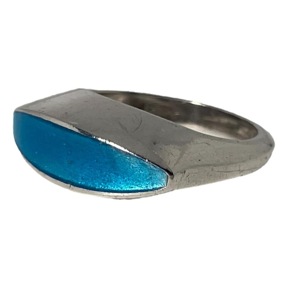 Photo of turquoise blue enamel ring displayed on a flat surface with the ring front facing to the bottom left corner. We are looking from a near eye level and the sleek design and smooth edges of the ring are clearly displayed.