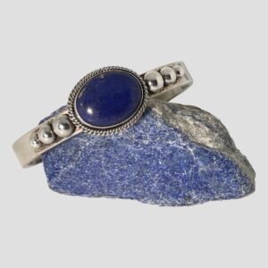 Main photo of Artie Yellowhorse cuff bracelet displayed on top of a small rock of lapis lazuli with the bracelet front forward facing. The bracelet is slightly angled down to the left so laid diagonally across the rock. It has a generous sized but not huge oval lapis cabochon with a double rope twist frame which is then stuck onto the cuff band, To either side of the lapis on the flat cuff band are 3 graduating silver balls starting with largest closest to the lapis. A very handsome chunky piece of quality Navajo jewellery.