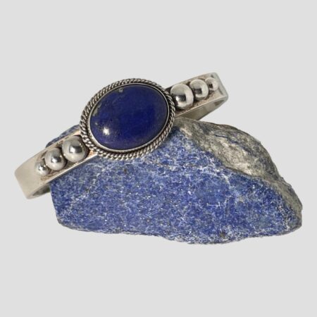 Main photo of Artie Yellowhorse cuff bracelet displayed on top of a small rock of lapis lazuli with the bracelet front forward facing. The bracelet is slightly angled down to the left so laid diagonally across the rock. It has a generous sized but not huge oval lapis cabochon with a double rope twist frame which is then stuck onto the cuff band, To either side of the lapis on the flat cuff band are 3 graduating silver balls starting with largest closest to the lapis. A very handsome chunky piece of quality Navajo jewellery.
