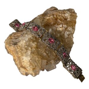 Main photo of Czech Deco paste bracelet displayed laid across a rock of natural river quartz with one end of the bracelet in top left and other in bottom right corner. The bracelet has 7 rectangular panels each with a pretty oval pink paste stone at the centre. The 4 claws holding the pink paste in place is brass tone. The panels are in a filigree style and mostly white metal with copper tones coming through. The rings holding the panels together (one at top and other on bottom edges) are also brass in tone.