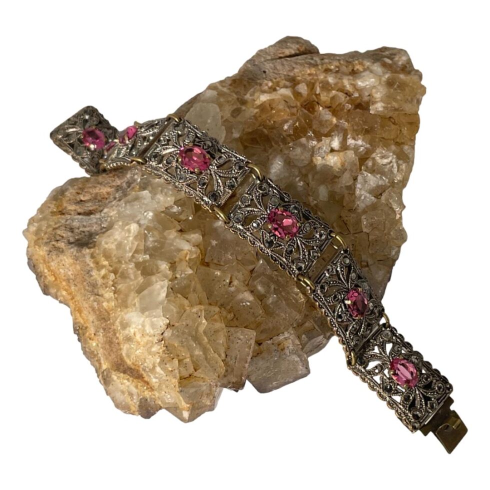 Main photo of Czech Deco paste bracelet displayed laid across a rock of natural river quartz with one end of the bracelet in top left and other in bottom right corner. The bracelet has 7 rectangular panels each with a pretty oval pink paste stone at the centre. The 4 claws holding the pink paste in place is brass tone. The panels are in a filigree style and mostly white metal with copper tones coming through. The rings holding the panels together (one at top and other on bottom edges) are also brass in tone.